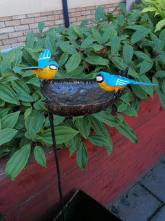 Bird bath recycled metal in the shape of a leaf with 2 bluetits birds, one with open wings & one with closed wings. Showing the leaf on a stick in an outdoor setting.