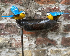 Bird bath recycled metal in the shape of a leaf with 2 bluetits birds, one with open wings & one with closed wings. Showing the leaf on a metal stick against a brick wall.