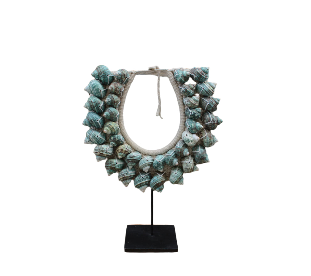 A handcrafted shell necklace stand featuring a variety of sea shells in white and periwinkle blue colors, displayed against a stone wall and wooden surface backdrop.