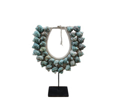 A handcrafted shell necklace stand featuring a variety of sea shells in white and periwinkle blue colors, displayed against a stone wall and wooden surface backdrop.