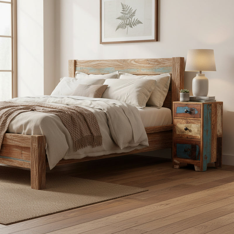 A small three-drawer wooden nightstand made from reclaimed wood with distressed blue and beige panels, rustic metal knobs, and small feet at the bottom, shown in a bright bedroom