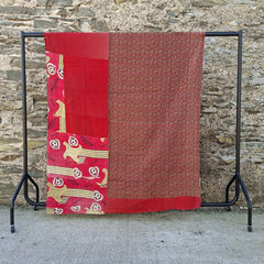 A red patchwork quilt with traditional South Asian motifs, draped over a black metal clothes rack against a rustic stone wall.