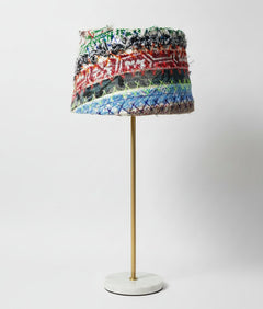 A table lamp with a cylindrical shade made of horizontal multicolored woven or embroidered bands, mounted on a slim gold metal stand with a round white base, photographed against a light gray studio background.