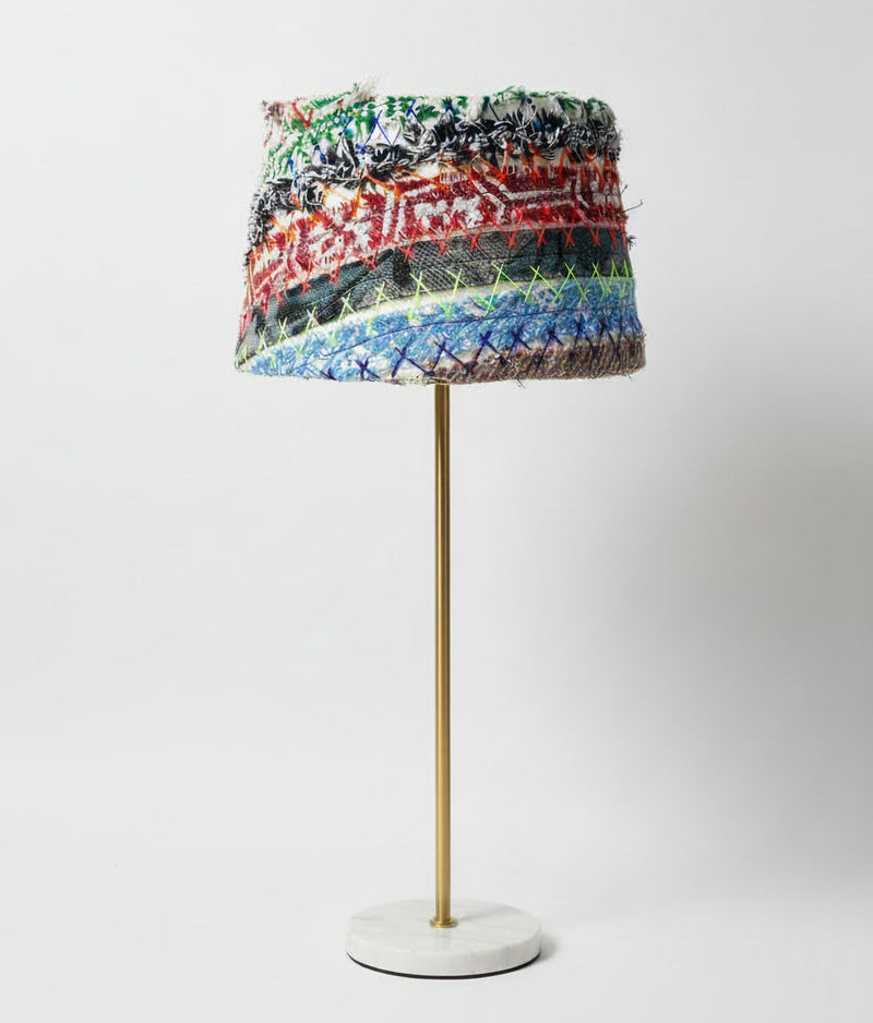 A table lamp with a cylindrical shade made of horizontal multicolored woven or embroidered bands, mounted on a slim gold metal stand with a round white base, photographed against a light gray studio background.