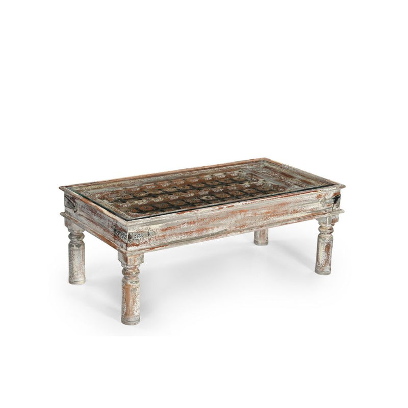 Wood coffee table with a distressed grey patina showing hints of natural wood. It has 4 round legs and a glass tp revealing a wood panels inspired by original shekawati indian door. Side view.