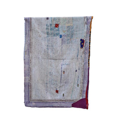 A vintage patchwork quilt with a light beige base, purple borders, and colorful geometric accents, displayed on a black metal stand against a rustic stone wall.